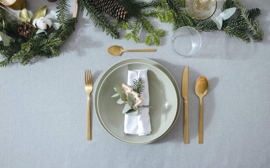 Imagem de uma mesa posta natalina  em tons de verde claro, talheres gold e guardanapo de tecido torcido com uma flor seca e biscoito amanteigado em formato de árvore de Natal.