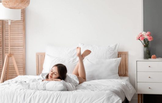 Foto que ilustra matéria sobre tipos de cama mostra uma jovem mulher deitada de bruços em uma cama, com o rosto voltado para o pé da cama e seus pés voltados para a cabeceira.