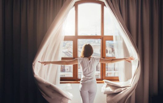 Foto que ilustra matéria sobre tipos de cortina mostra uma mulher de costas abrindo cortinas e olhando por uma janela em um dia de sol.