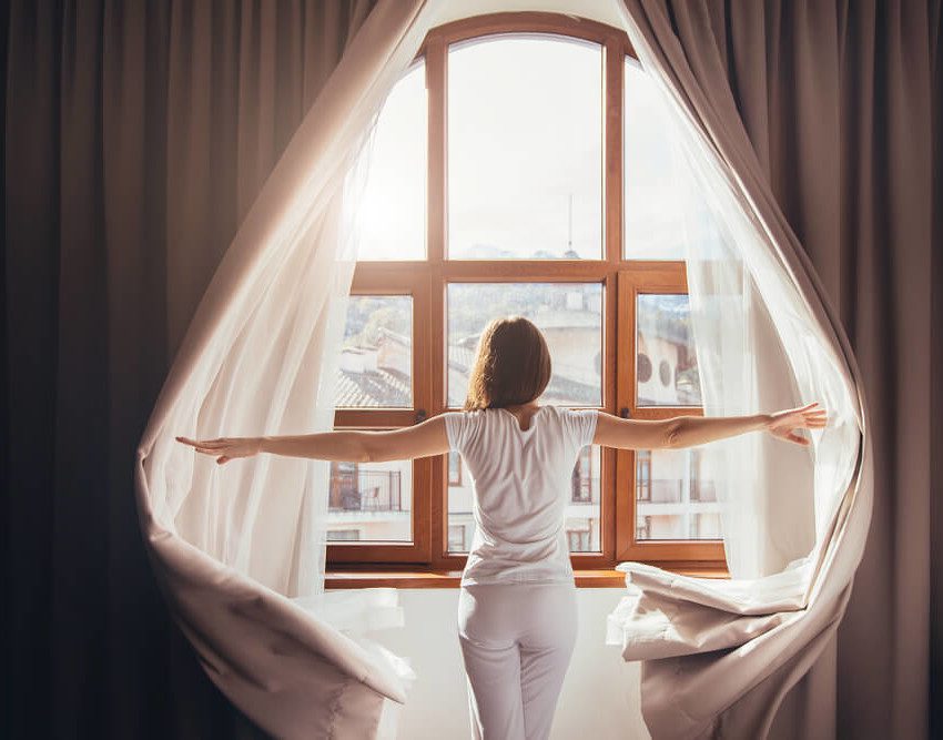 Foto que ilustra matéria sobre tipos de cortina mostra uma mulher de costas abrindo cortinas e olhando por uma janela em um dia de sol.