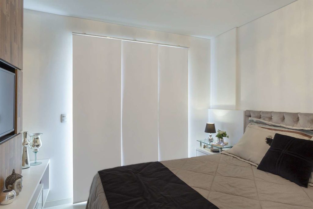 Foto que ilustra matéria sobre tipos de cortina mostra um quarto com cama de casal feita. Ao fundo, há uma cortina do tipo painel branca com três lâminas verticais cobrindo uma janela.