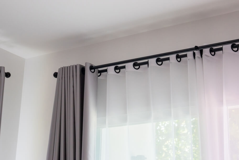 Foto que ilustra matéria sobre tipos de cortina mostra uma janela com duas cortinas, um blecaute cinza e uma de voal branca, presas em dois varões pretos.