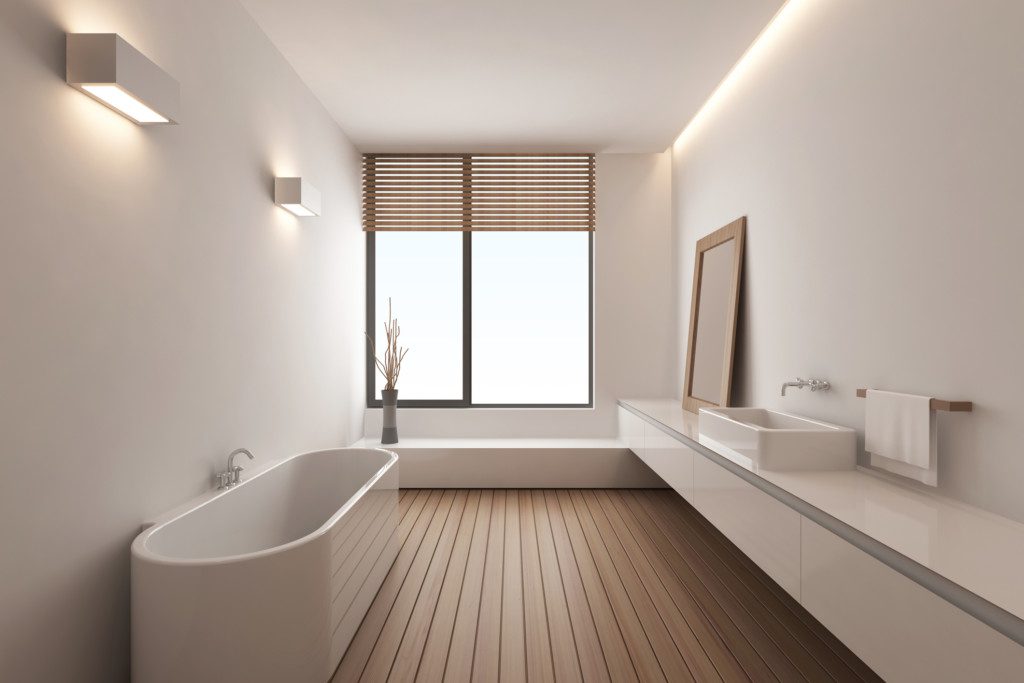 Foto de um banheiro com uma janela ao fundo, por onde entra a luz natural. Há também um piso de madeira, uma banheira oval e uma pia com armário em linhas retas, além de um espelho em formato quadrado.