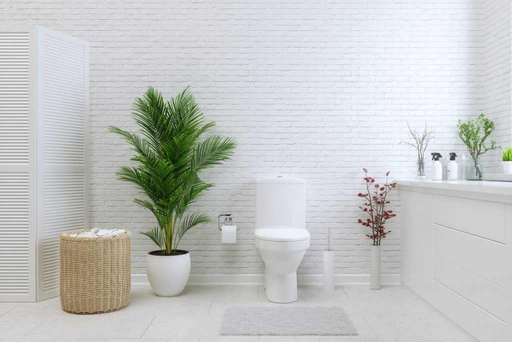 Foto de um banheiro com parede em tijolos brancos que tem plantas complementando a decoração. Há uma planta maior em um vaso branco, outra menor com galhos coloridos e duas pequenas na pia. Tem também um cesto de roupas, um vaso e uma pia.
