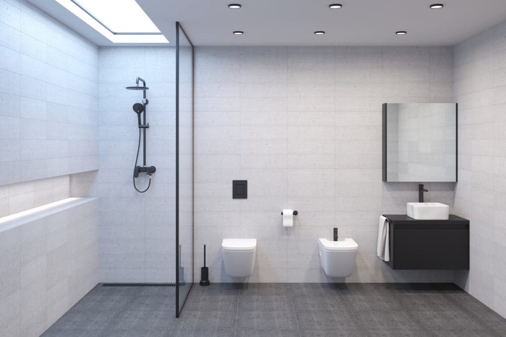 Foto de um banheiro com chuveiro minimalista moderno e iluminado com uma lâmpada de luz do dia no teto para criar a ilusão de uma janela. Há também ferragens escuras para criar contraste com paredes claras. Além de bidê e vaso sanitário branco.