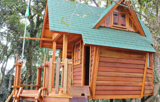 Foto que ilustra matéria sobre casa na árvore simples mostra uma pequena casa de madeira com telhado verde apoiada no tronco de uma árvore.