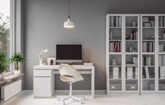 Foto de um escritório com escrivaninha, cadeira, lustre de teto e estante em cor branca. A parede e o computador complementam na cor cinza.