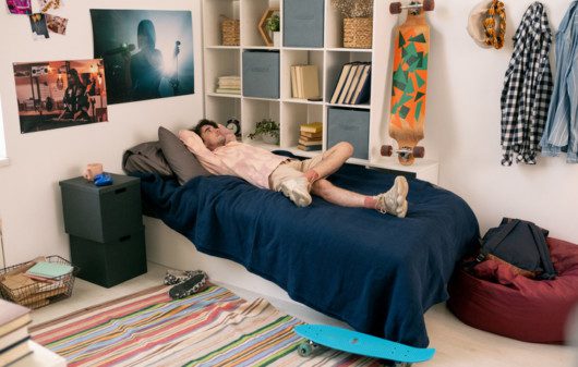 Foto que ilustra matéria sobre quarto geek mostra um homem jovem deitado na cama com posteres nas paredes e um skate pendurado