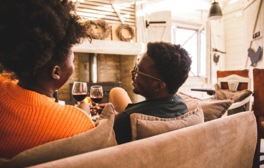 Vista traseira de um jovem casal relaxando no sofá em uma sala de estar rústica, tendo momentos de união e brindando com taças de vinho tinto.
