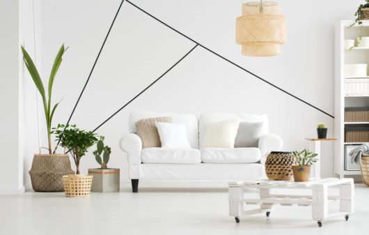 Foto que ilustra matéria sobre parede geométrica mostra uma sala de estar minimalista com um sofá, uma mesa de centro e uma estante de livros brancos. Na parede atrás, há uma pintura apenas com o contorno de triângulos.