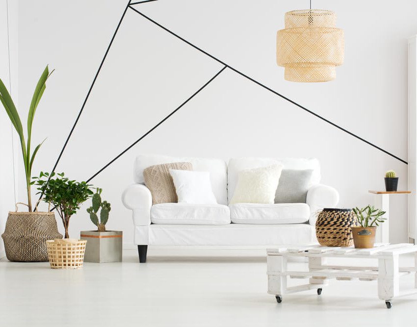 Foto que ilustra matéria sobre parede geométrica mostra uma sala de estar minimalista com um sofá, uma mesa de centro e uma estante de livros brancos. Na parede atrás, há uma pintura apenas com o contorno de triângulos.
