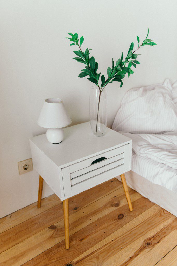 Mesa de cabeceira branca com pés palito em madeira, abajur branco e vaso de planta minimalista.