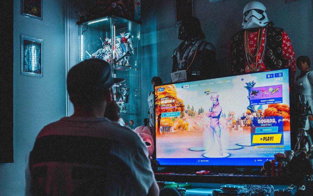 Na imagem tem um homem sentado no tapete, jogando vídeo game. Atrás da TV há diferentes bonecos e itens nerds.
