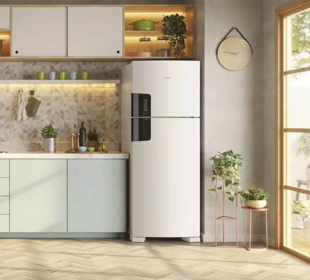 Foto que ilustra matéria sobre quanto custa mobiliar um apartamento mostra uma cozinha com uma geladeira branca posicionada entre uma pilastra e a bancada da pia com armários embutidos abaixo. Acima da geladeira há mais armários e prateleiras