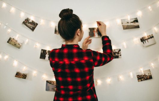 Foto que ilustra matéria sobre quarto tumblr mostra uma mulher pendurando fotos em um varal de luzes em uma parede