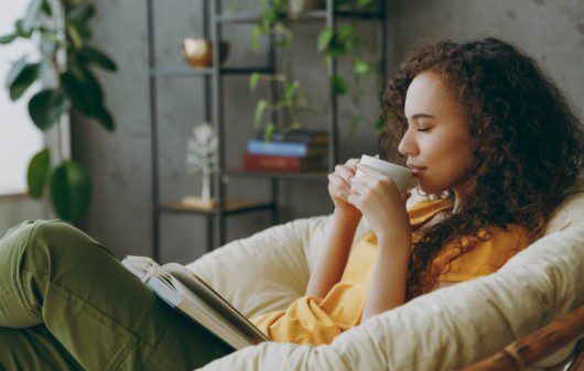 Foto que ilustra matéria sobre sala de descanso mostra uma jovem mulher sentada em uma poltrona com um livro no colo e tomando uma bebida em uma xícara com os olhos fechados