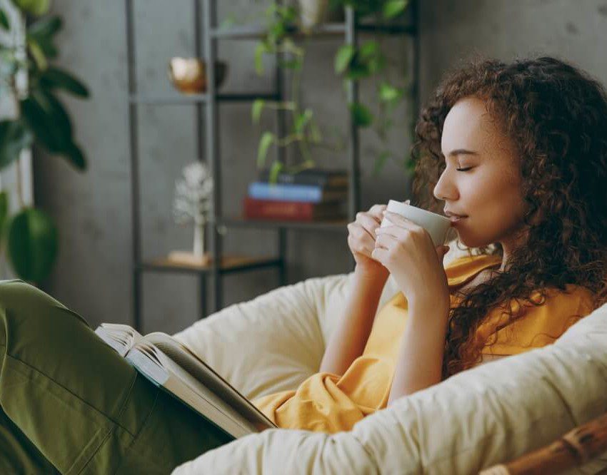 Foto que ilustra matéria sobre sala de descanso mostra uma jovem mulher sentada em uma poltrona com um livro no colo e tomando uma bebida em uma xícara com os olhos fechados