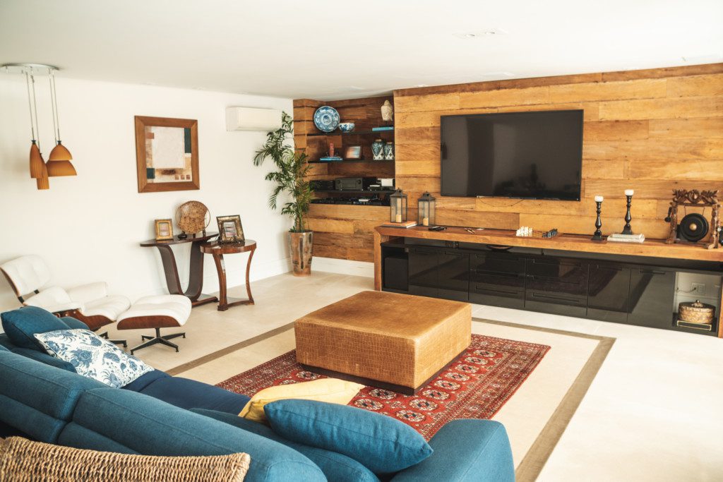 Foto de uma sala de estar rústica com um painel e rack de madeira ocupando toda uma parede. Há também elementos decorativos como: sofá, lustre em tom de terroso, mesa de centro, tapete, quadro, plantas e outros.