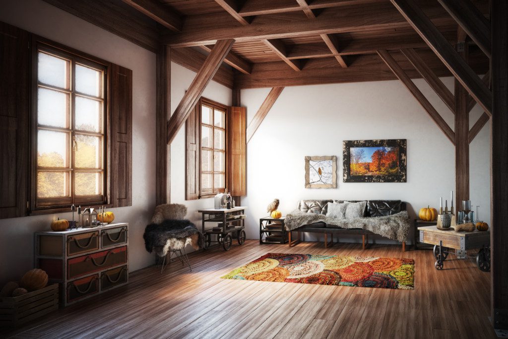 Foto de uma sala de estar rústica com diferentes elementos decorativos como: tapete, almofadas, manta e outros. A sala é feita em piso e vigas de madeira. Há nela um sofá em destaque.