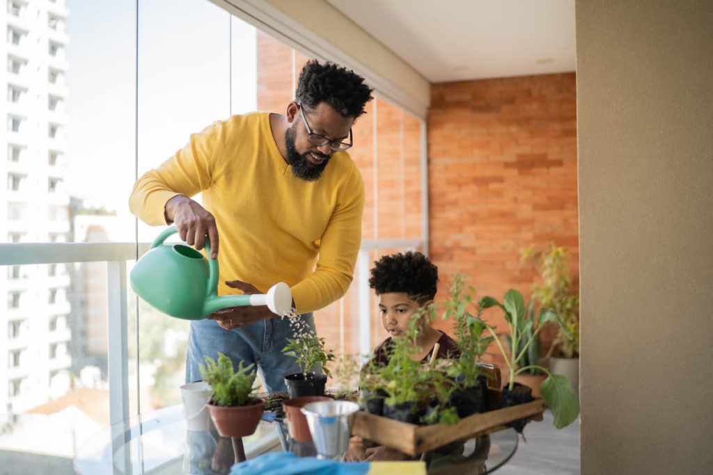 Pai e filho negros regam plantas na varanda.