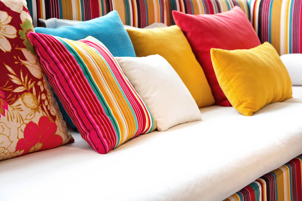 Foto que ilustra matéria sobre varanda tropical mostra um sofá com listras coloridos com diversas almofadas também de diferentes cores.