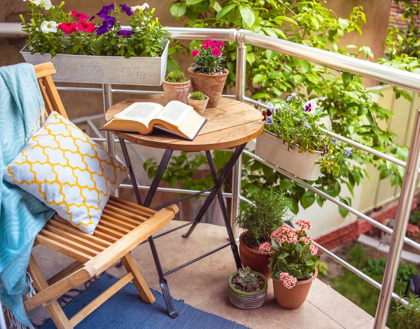 Foto que ilustra matéria sobre varanda tropical mostra uma varanda com uma cadeira e uma mesinha de madeira, cercadas de plantas. Na mesinha há um livro aberto e na cadeira há uma almofada e uma manta.