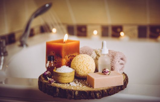 Imagem de velas aromáticas, toalhas, esponja, escova e óleos corporais para um banho relaxante.