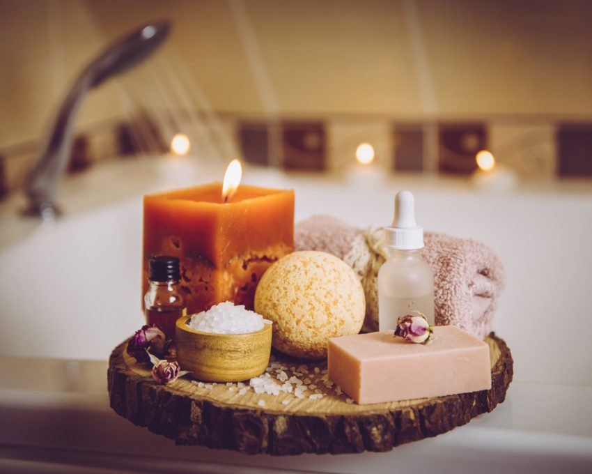 Imagem de velas aromáticas, toalhas, esponja, escova e óleos corporais para um banho relaxante.