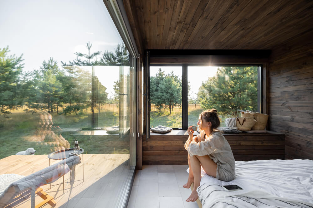 Foto que ilustra matéria sobre design sensorial mostra uma mulher sentada na beira de uma cama co uma xícara de café em um quarto com uma grande parede de vidro que dá para uma varanda e uma grande área verde.