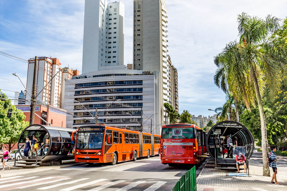Foto que ilustra matéria sobre transporte público em Curitiba mostra duas estações do BRT, cada uma com um ônibus biarticulado ao lado, virados para direções opostas.