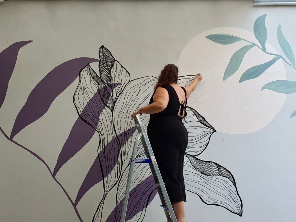 Mulher em uma escada desenhando flores em uma parede. 