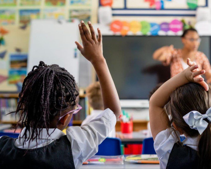 Uma menina, em sala de aula, levanta a mão para tirar dúvidas com a professora.