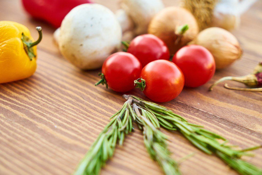 Imagem de um alecrim em cima de uma mesa ao lado de cebolas,pimentões e tomates
