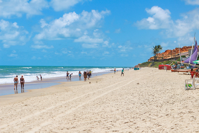 Trecho da praia do Morro Branco mostra pessoas no mar, jangadas na areia, mesas e cadeiras de plástico. 