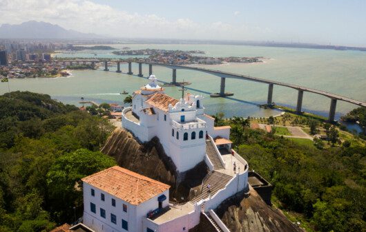 Foto mostra o Convento da Penha visto de cima, no alto de um morro, com a baía e a cidade de Vila Velha ao fundo.