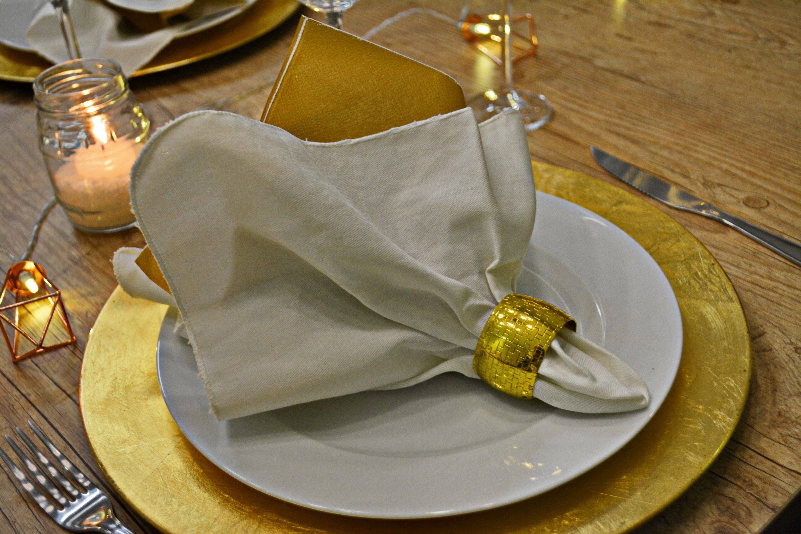 Sousplat em alumínio dourado, prato e guardanapo de tecido branco com argola dourada. 
