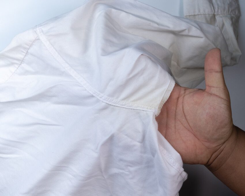 Imagem de uma mão segurando uma camiseta branca com mancha de desodorante amarelada.