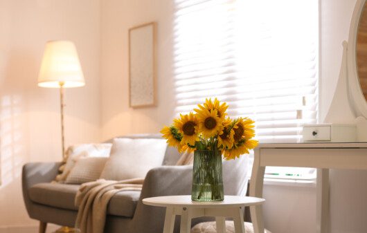 Imagem de uma sala de estar em tons neutros, com um vaso de flor de girassóis em cima da mesa lateral.
