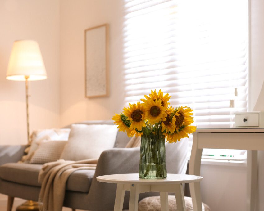Imagem de uma sala de estar em tons neutros, com um vaso de flor de girassóis em cima da mesa lateral.