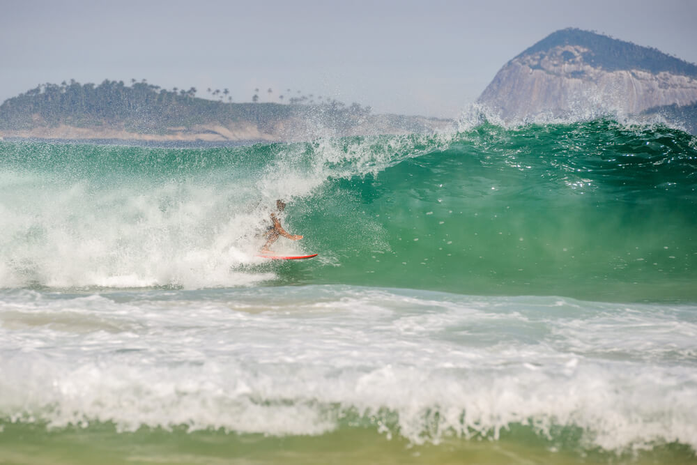 Foto que ilustra matéria sobre ilha no Rio de Janeiro mostra um surfista em ação pegando uma onda em primeiro plano. E ao fundo aparece uma grande rocha que faz parte das Ilhas Cagarras.