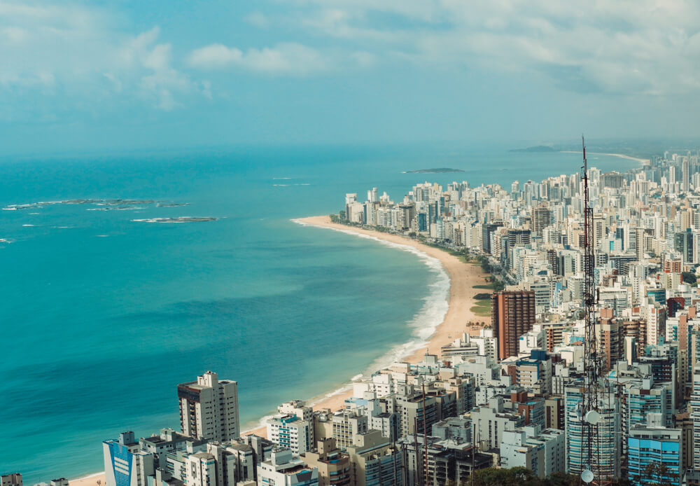 Foto que ilustra matéria sobre Praias de Vila Velha mostra uma visão do alto da Praia da Costa, com o mar à esquerda, a faixa de areia no meio e prédios à direita da imagem. 