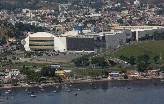 Foto mostra uma visão do alto e de longe do São Gonçalo Shopping, com uma parte da Baía de Guanabara aparecendo em primeiro plano, o shopping no Centro e a cidade atrás.