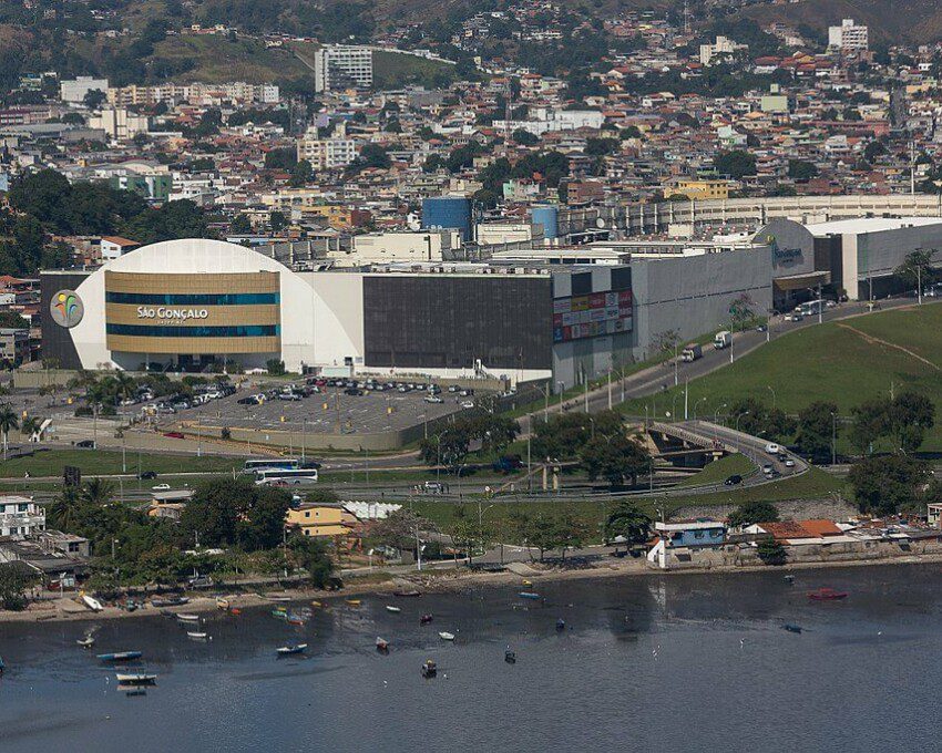 Foto mostra uma visão do alto e de longe do São Gonçalo Shopping, com uma parte da Baía de Guanabara aparecendo em primeiro plano, o shopping no Centro e a cidade atrás.
