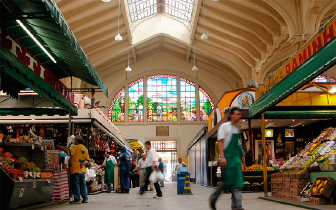 Imagem interna do primeiro andar do Mercado Municipal de SP com diversas pessoas passando por lá.