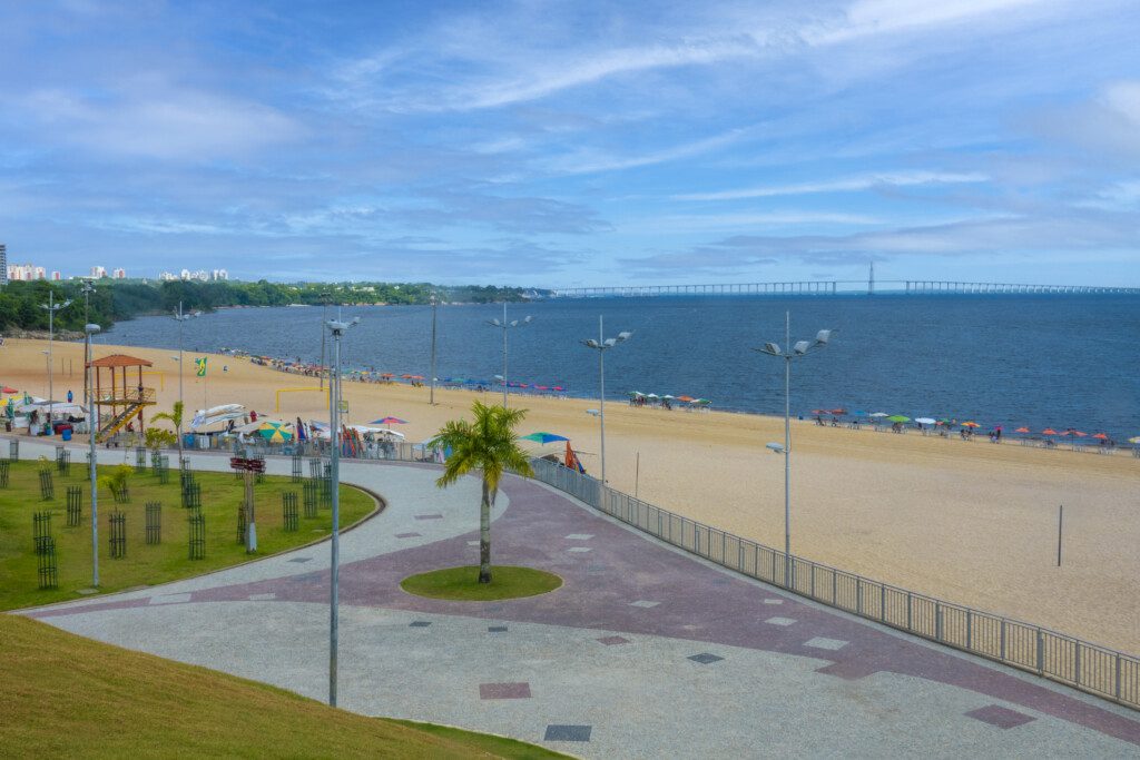 A foto mostra a parte da orla da praia que tem calçadão e barracas de comida e bebida. Há também a areia e o mar, além de árvores. 