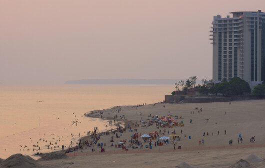 Foto da Praia de Ponta Negra durante o pôr do sol. Há pessoas na areia e prédios ao fundo.
