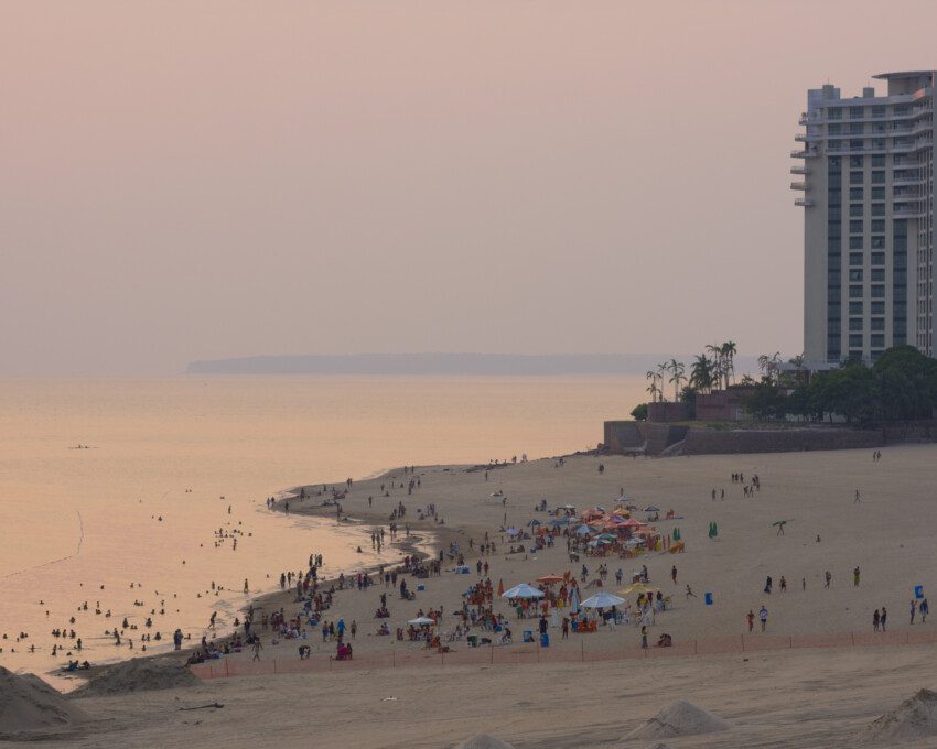 Foto da Praia de Ponta Negra durante o pôr do sol. Há pessoas na areia e prédios ao fundo.