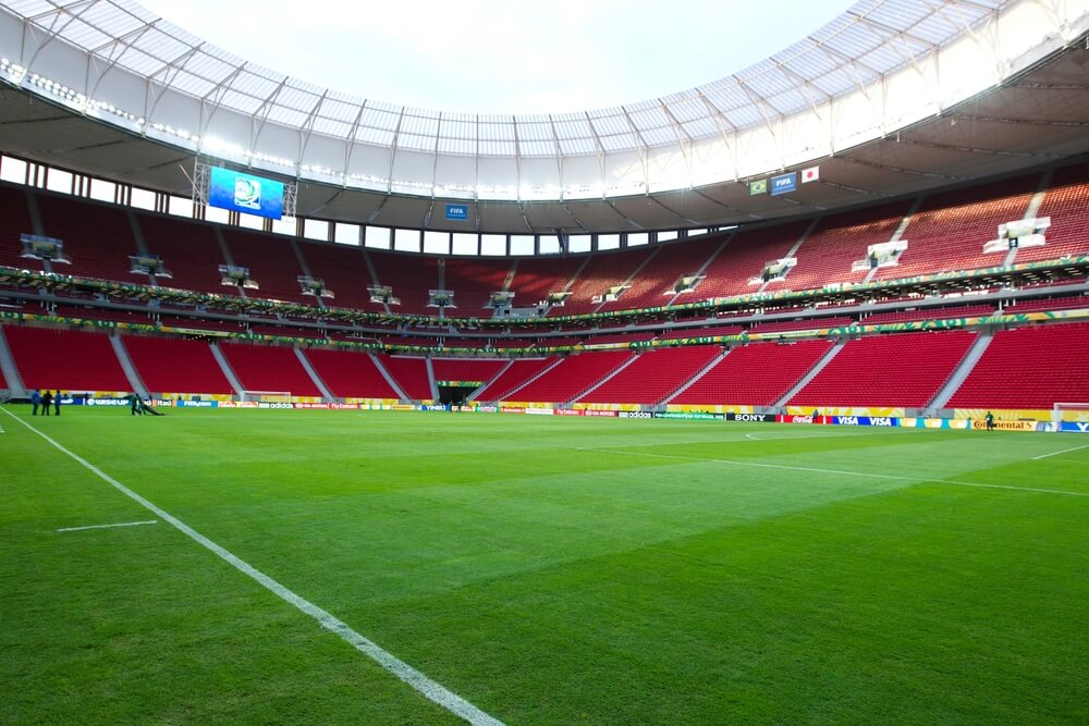 Foto que ilustra matéria sobre o Estádio Mané Garrincha mostra o interior da arena, com seu gramado e as arquibancadas com cadeiras vermelhas.