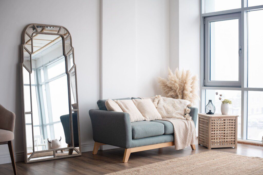 sala de estar com espelho de chão, sofá verde com almofadas e mantas, mesinha lateral e plantas.