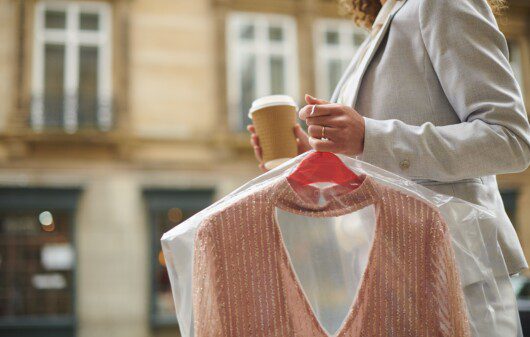 Imagem de uma mulher segurando um café com uma mão e não outra uma peça de roupa recém lavada na lavanderia.